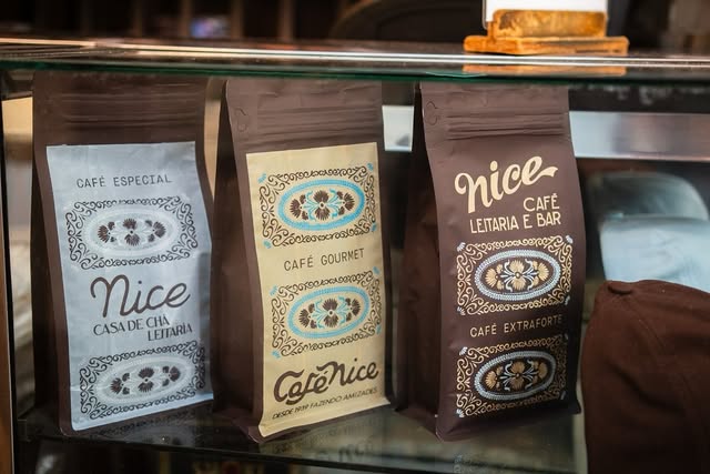 Três pacotes de café do Nice em uma vitrine de vidro: um azul-acinzentado, um cor de creme e um marrom, todos com estampas em ovais ornamentadas.