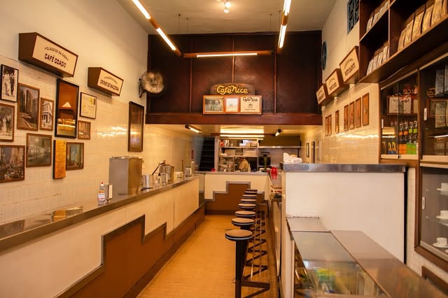 Interior do Café Nice após a reforma: o corredor do balcão de inox, com bancos altos, azulejos claros, quadros e o letreiro Café Nice ao fundo.