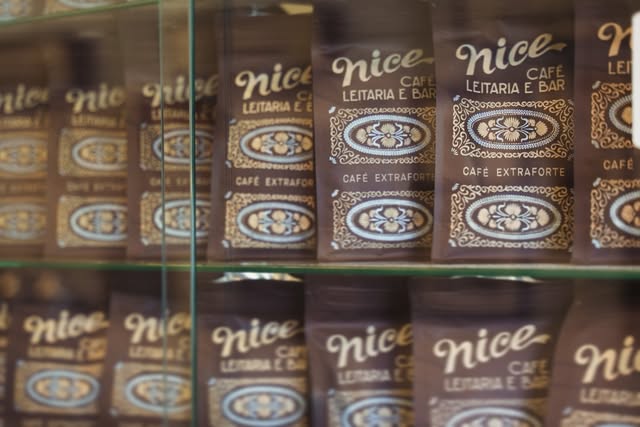 Pacotes de café Nice, Café, Leitaria e Bar, empilhados atrás do vidro de uma vitrine, em marrom e dourado.