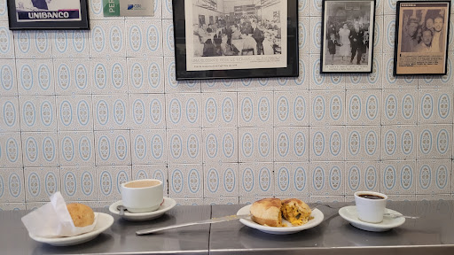 Parede de azulejos azul-acinzentados com quadros antigos emoldurados acima do balcão de inox, onde há xícaras de café e um pão de queijo.