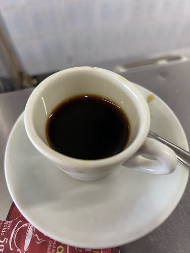 Cafezinho coado em xícara pequena de porcelana branca sobre pires, no balcão de inox.