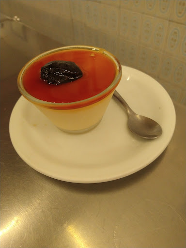 Creme de maizena com calda de caramelo e uma ameixa por cima, em copinho de vidro sobre um pires branco, no balcão de inox.
