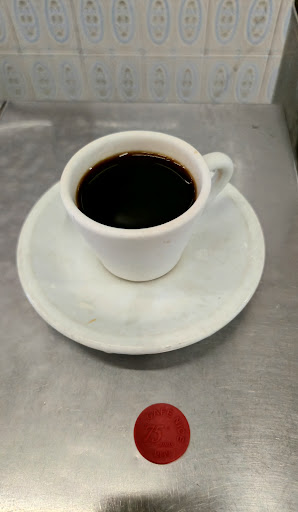 Cafezinho preto em xícara branca sobre pires, no balcão de inox, com uma ficha vermelha ao lado.