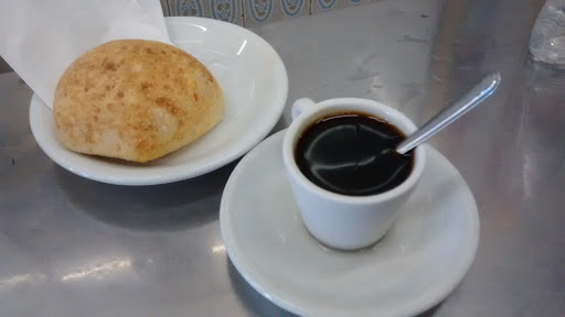 Balcão de inox com pão de queijo, um sanduíche e duas xícaras de café diante da parede de azulejos e de quadros antigos.