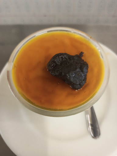 Creme de maizena com ameixa visto de cima, coberto de calda dourada, em copo de vidro sobre pires branco.