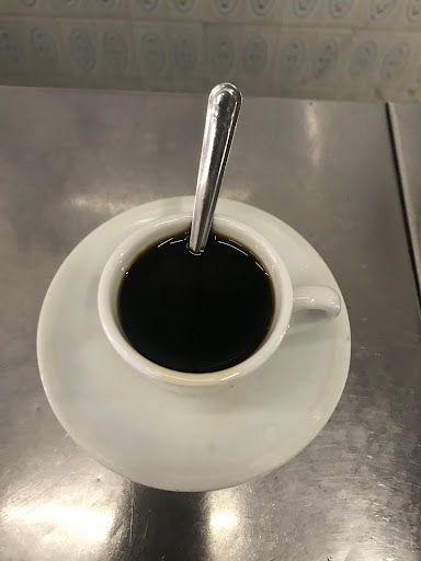 Cafezinho preto em xícara branca com colher, visto de cima, sobre o pires e o balcão de inox.
