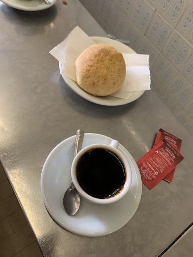 Xícara de café preto com colher sobre um pires e, ao fundo, um pão de queijo em pratinho com guardanapo, sobre o balcão de inox ao lado da parede de azulejos.