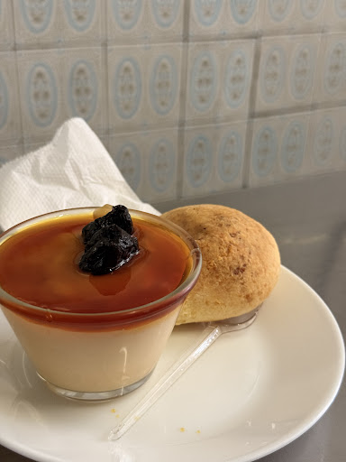 Creme de maizena com ameixa ao lado de um pão de queijo, em prato branco, diante de azulejos azul-acinzentados.