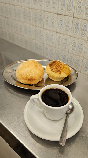 Dois pães de queijo em uma bandeja de inox e um cafezinho preto em xícara branca com colher, diante da parede de azulejos.