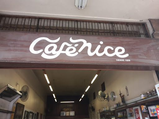 Letreiro de madeira da fachada com o nome Café Nice em letras brancas cursivas e a inscrição Desde 1939, com a entrada do café logo abaixo.